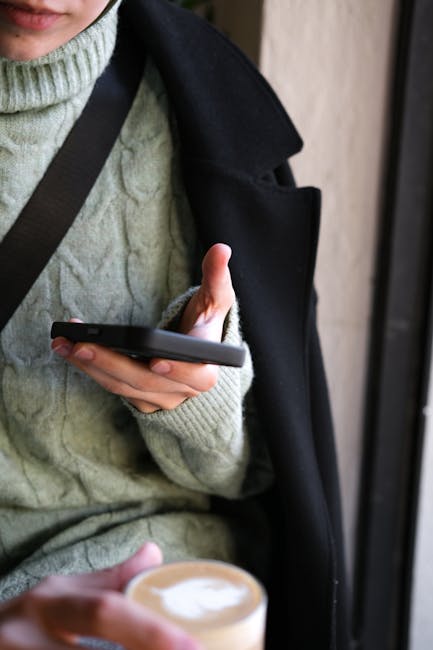 Adult in cozy green sweater uses smartphone while holding coffee indoors.